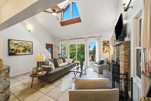Un salon spacieux avec une cheminée en pierre, une grande fenêtre donnant sur un paysage verdoyant et un ventilateur de plafond.