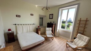 A bedroom with a bed, two chairs, a wooden dresser, a nightstand, a lamp, a window, and a ceiling light fixture. - Apartment in a beautiful house in the countryside near the ocean. (Plévenon)