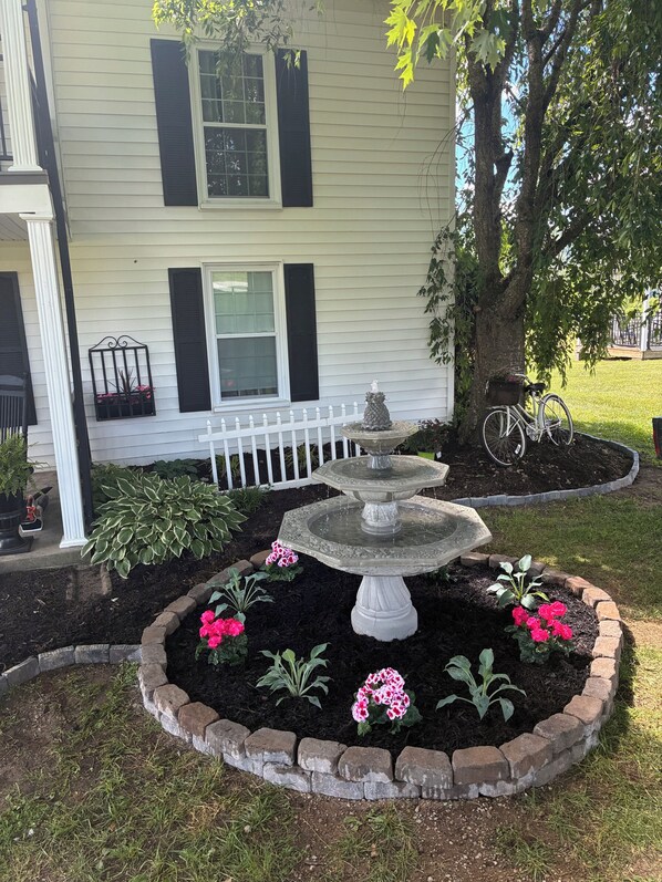 A house with a white exterior, black shutters, and a front yard with a stone fountain, flower beds, and a white picket fence.