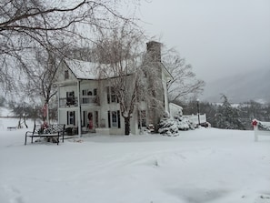 Una casa de dos pisos con un balcón, rodeada de árboles cubiertos de nieve y una valla de piquetes blancos.