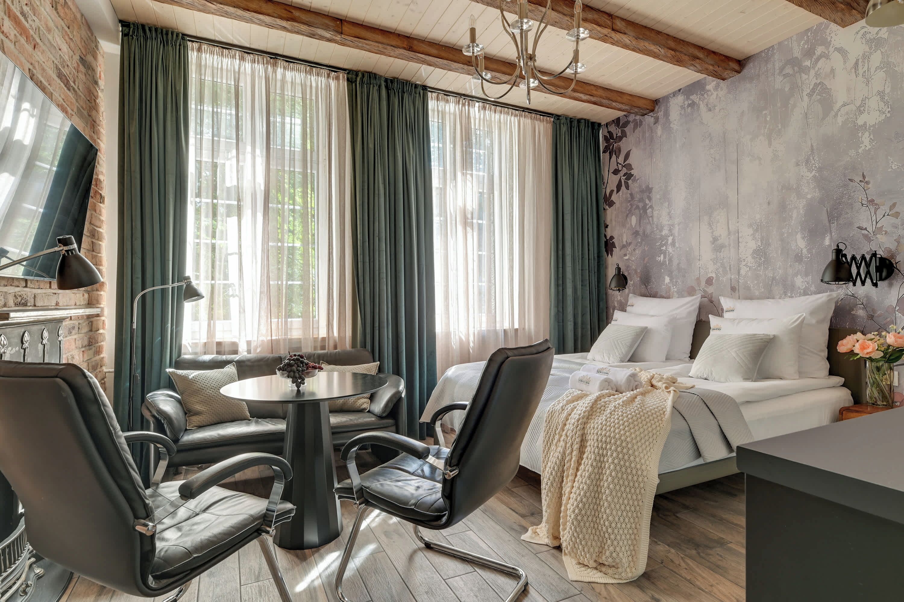 A room with a bed, a dining table with chairs, a chandelier, and a view through large windows with curtains.