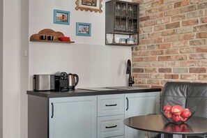 A kitchen with a brick wall, a coffee maker, a kettle, a cabinet with glassware, and a table with a bowl of apples. - Charm Atelier Apartment 3A | Old Town Ducha Street (Gdańsk)