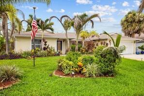 A residential property with a well-maintained lawn, a variety of tropical plants, and a single-story house with a garage. - Right Direction Rentals Tropical Haven Retreat (Port St Lucie)