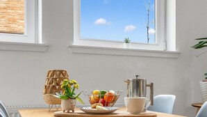 A dining area with a wooden table, blue chairs, a fruit basket, a coffee pot, and a view of the sky through large windows. - Edinburgh CITY Centre 2BR Apartment (Edinburgh)