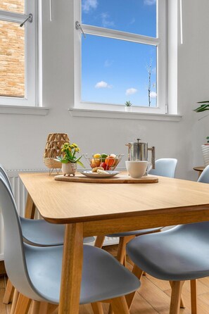 A dining area with a wooden table, blue chairs, a fruit basket, a coffee pot, and a view of the sky through large windows. - Edinburgh CITY Centre 2BR Apartment (Edinburgh)