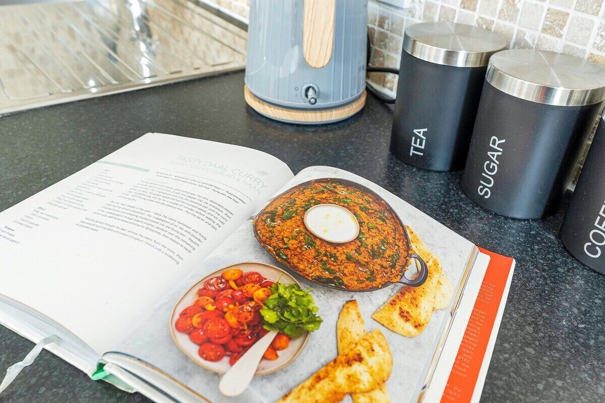 A cookbook open on a kitchen counter with a recipe for a dish, a bowl of salad, and two containers labeled 'TEA' and 'SUGAR'.