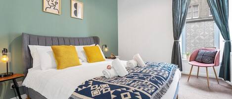 A bedroom with a bed, pillows, a blanket, a chair, and a nightstand.