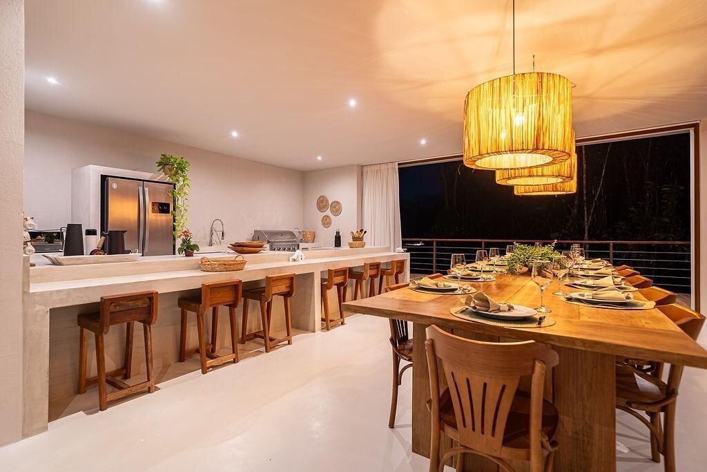 A modern kitchen with a dining area, featuring a long wooden table set for six, a built-in island with bar stools, and a large window providing a view.