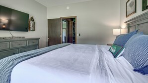 A bedroom with a large bed, a flat-screen TV, a wooden dresser, and a door leading to another room. - New! Riverfront Trout Fishing White River Ozarks (Cotter)