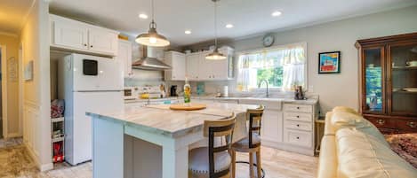 A kitchen with white cabinets, a central island, bar stools, pendant lighting, and a dining area with a sofa.