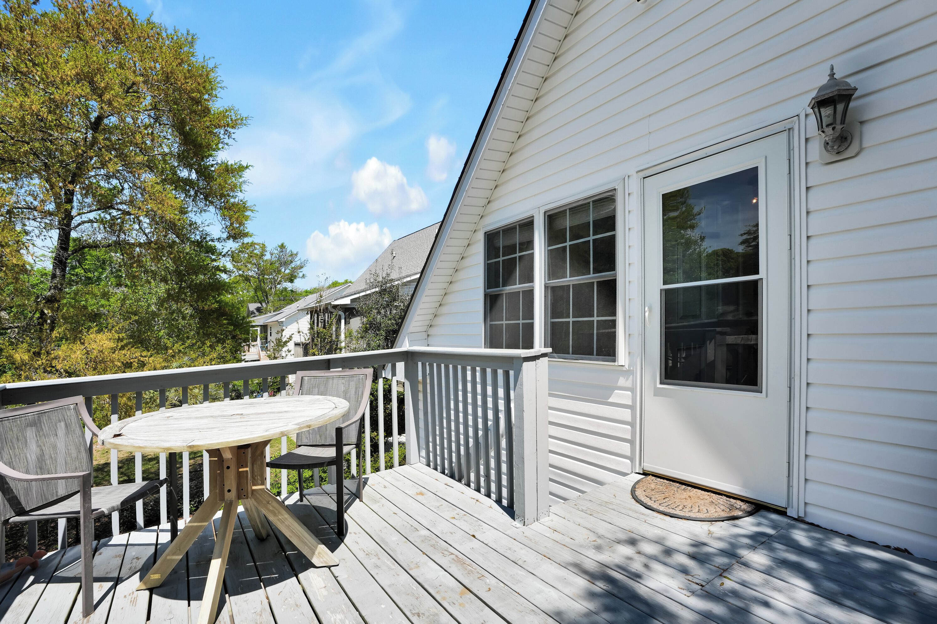 A deck with a round table and chairs, a white exterior wall, and a large window.