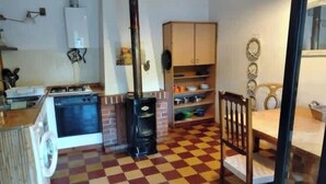 A kitchen with a stove, oven, and a black stove pipe. There is a checkered floor, a wooden chair, and a cabinet with dishes. - Traditional Country Home (São Brás de Alportel)