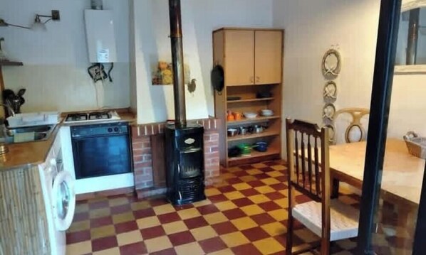 A kitchen with a stove, oven, and a black stove pipe. There is a checkered floor, a wooden chair, and a cabinet with dishes. - Traditional Country Home (São Brás de Alportel)