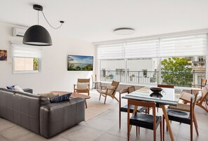 A modern living room with a grey sofa, a dining table with chairs, and a large window with a view of the sea. - Spacious Quiet 2BR & Parking City Center by FeelHome (Tel Aviv - Jaffa)