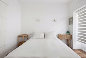 A minimalist bedroom with a large bed, a bedside table with a lamp, and a small side table. - Spacious Quiet 2BR & Parking City Center by FeelHome (Tel Aviv - Jaffa)