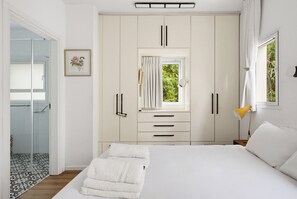 Un dormitorio con cama, armario, ventana con cortinas, lámpara de noche y puerta que da a un baño.