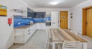 A modern kitchen with white cabinets, a blue backsplash, and a wooden dining table with chairs. - Modern 2BR Apartment w/ Balcony, St Paul`s Bay (San Pawl il-Baħar)