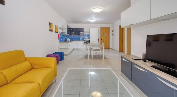 A modern living room with a yellow sofa, a dining area with a table and chairs, a kitchen with cabinets and appliances, and a TV on a wooden stand. - Modern 2BR Apartment w/ Balcony, St Paul`s Bay (San Pawl il-Baħar)