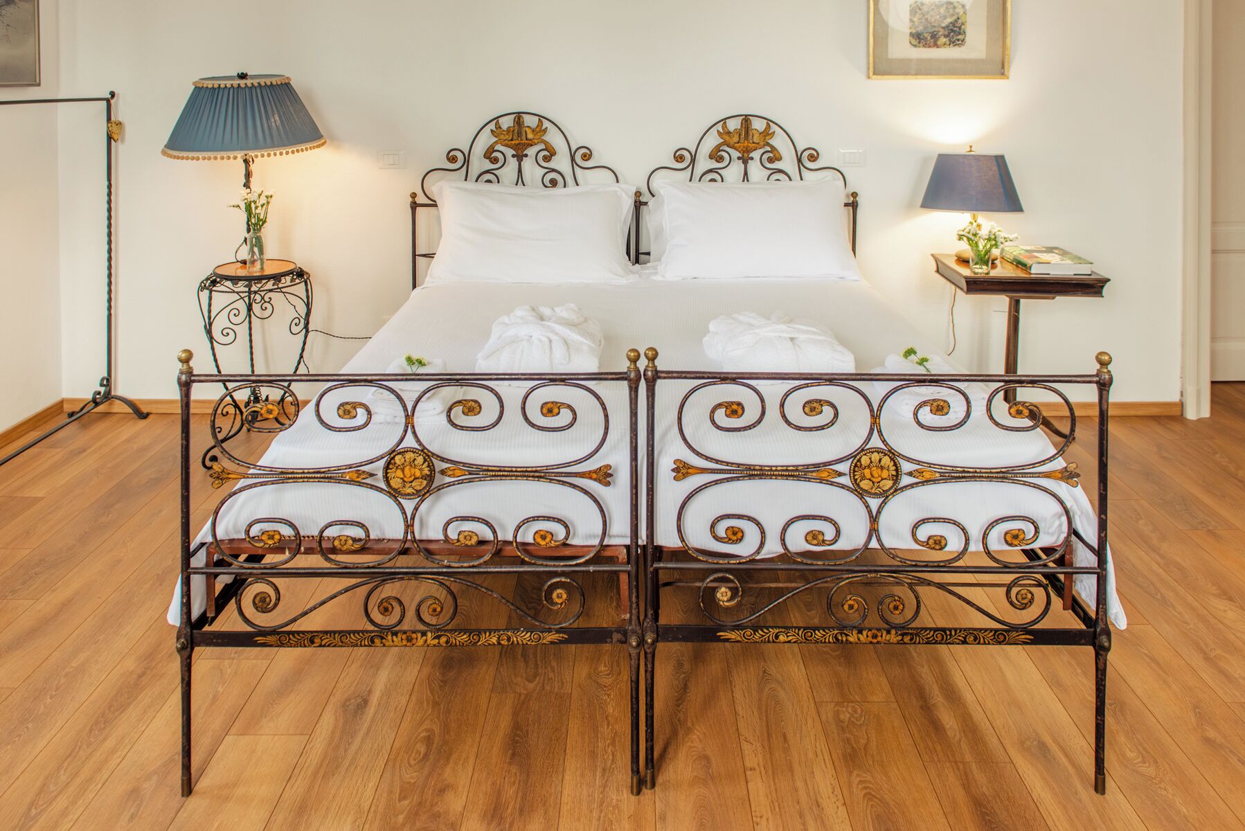 A double bed with ornate metal headboard and footboard, a bedside table with a lamp, and a framed picture on the wall.