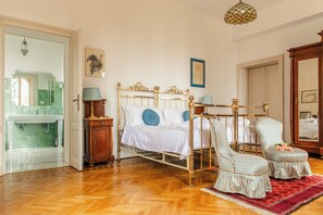 A bedroom with a bed, two chairs, a nightstand, a lamp, a mirror, and a door leading to a bathroom. - Villa Evelina (Travedona-Monate)