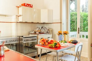 A kitchen with a stove, oven, and cabinets, a dining table with vegetables, and a window with a view of greenery. - Villa Evelina (Travedona-Monate)