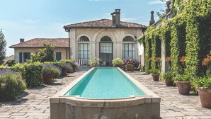 A swimming pool with a stone border, surrounded by potted plants and a building with arched windows. - Villa La Limonera (Casciago)