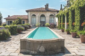 A swimming pool with a stone border, surrounded by potted plants and a building with arched windows. - Villa La Limonera (Casciago)