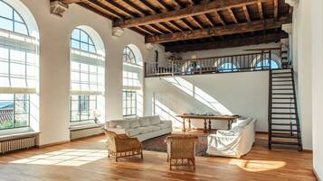 Un loft espacioso con techos altos, vigas de madera a la vista, un sofá grande, sillas de mimbre y vistas del cielo a través de ventanas arqueadas.