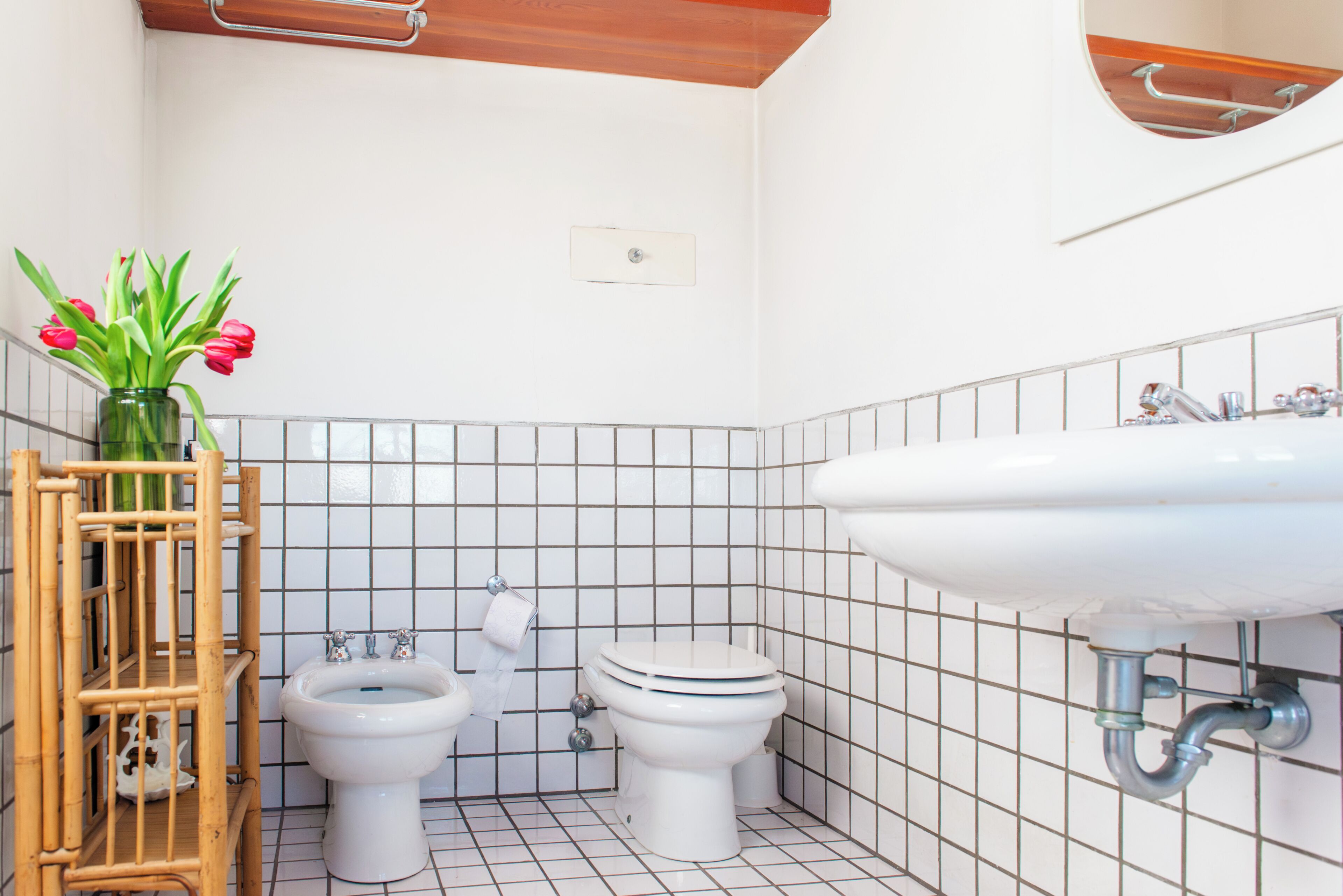 Ein Badezimmer mit einem weißen Waschbecken, zwei Toiletten, einem Bidet und einer Bambusablage mit Blumen in einer Vase.