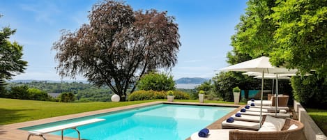 A swimming pool with lounge chairs and a pergola, surrounded by greenery and a view of the landscape.