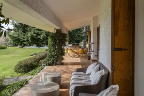 A covered outdoor seating area with a sofa, a table, and chairs, a grill, and a view of a well-maintained lawn and trees. - Villa Serenity (Besozzo)