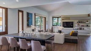 Eine moderne Küche mit einer großen Kochinsel, einem Essbereich mit langem Tisch und Stühlen sowie einem Wohnbereich mit Sofa und Fernseher.