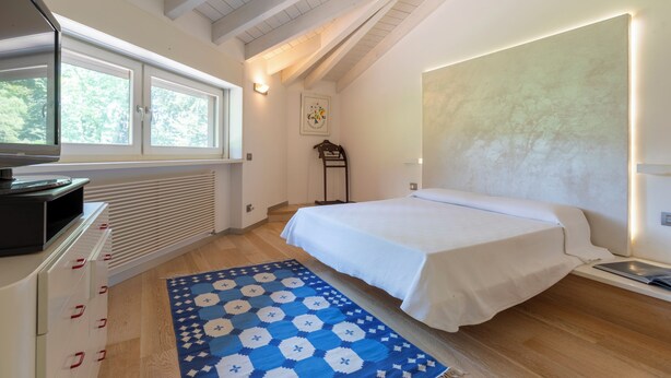 A bedroom with a bed, a radiator, a window, a desk, and a patterned rug. - Villa La Fortezza (Dormelletto)