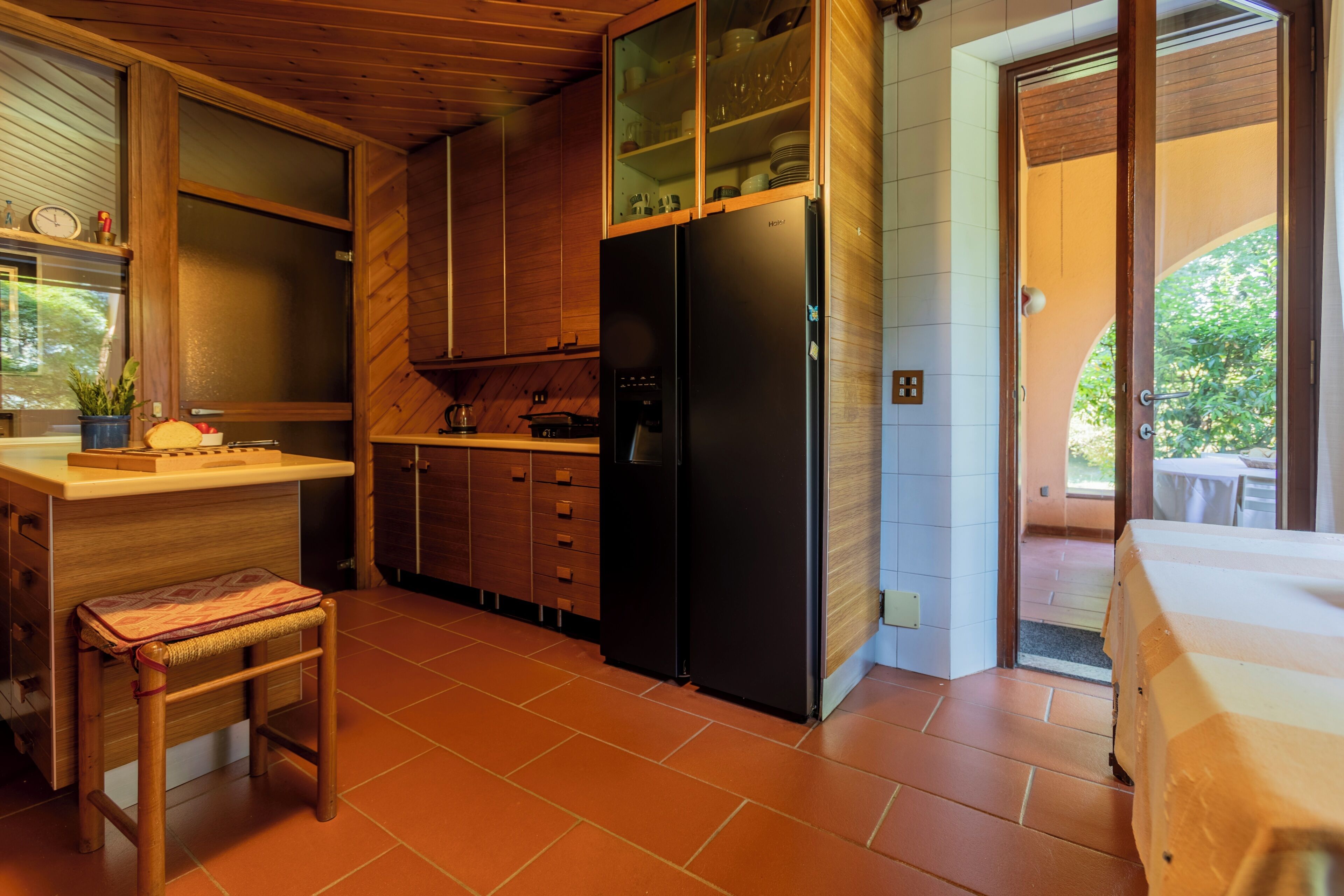 Une cuisine compacte avec des meubles de rangement en bois, un réfrigérateur noir et un petit tabouret.