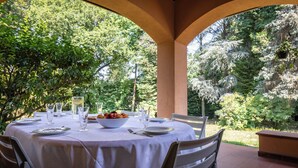 An outdoor dining area with a round table set for two, surrounded by chairs, with a view of trees and greenery. - Villa La Fortezza (Dormelletto)