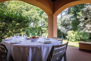 An outdoor dining area with a round table set for two, surrounded by chairs, with a view of trees and greenery. - Villa La Fortezza (Dormelletto)