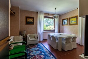 A room with a dining table, chairs, a sofa, a green chair, a patterned rug, a lamp, and framed pictures on the wall.