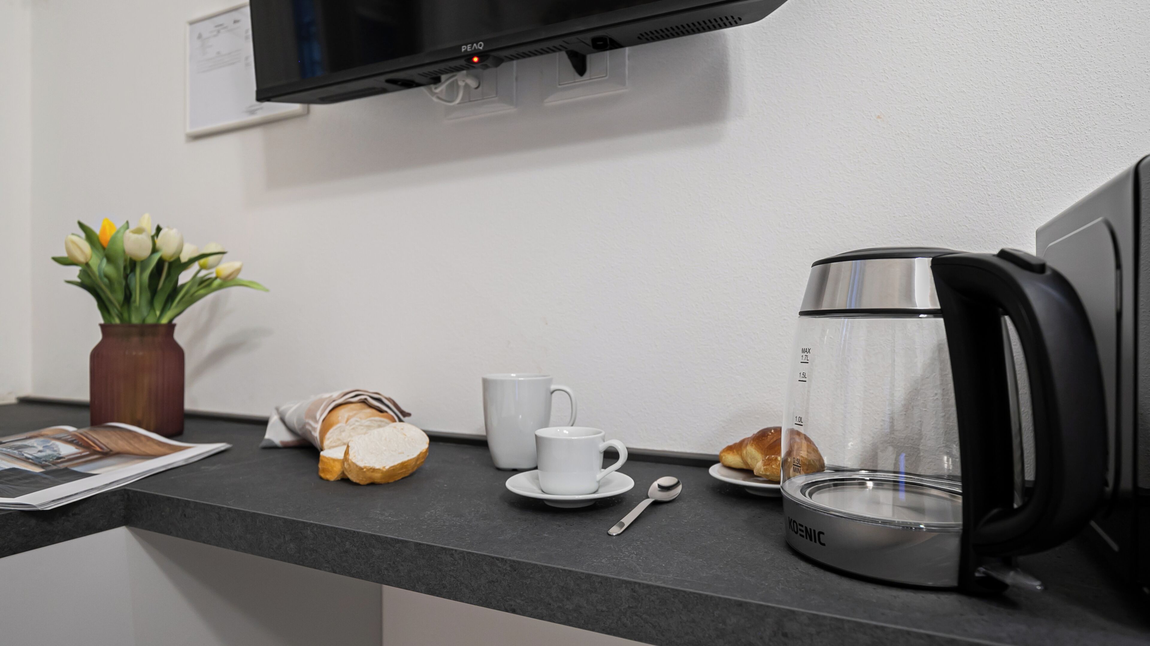 Een kitchenette met een koffiezetapparaat, een kop, een bord met croissant, een lepel en een vaas met bloemen.