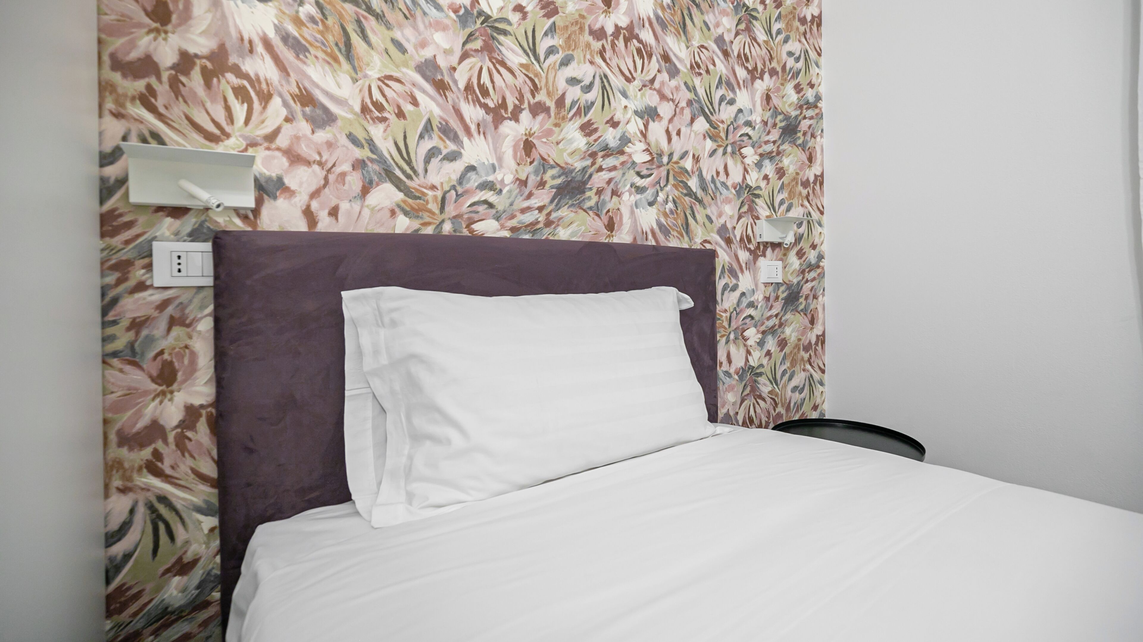 A bed with white bedding, a purple headboard, and a floral wallpaper pattern.