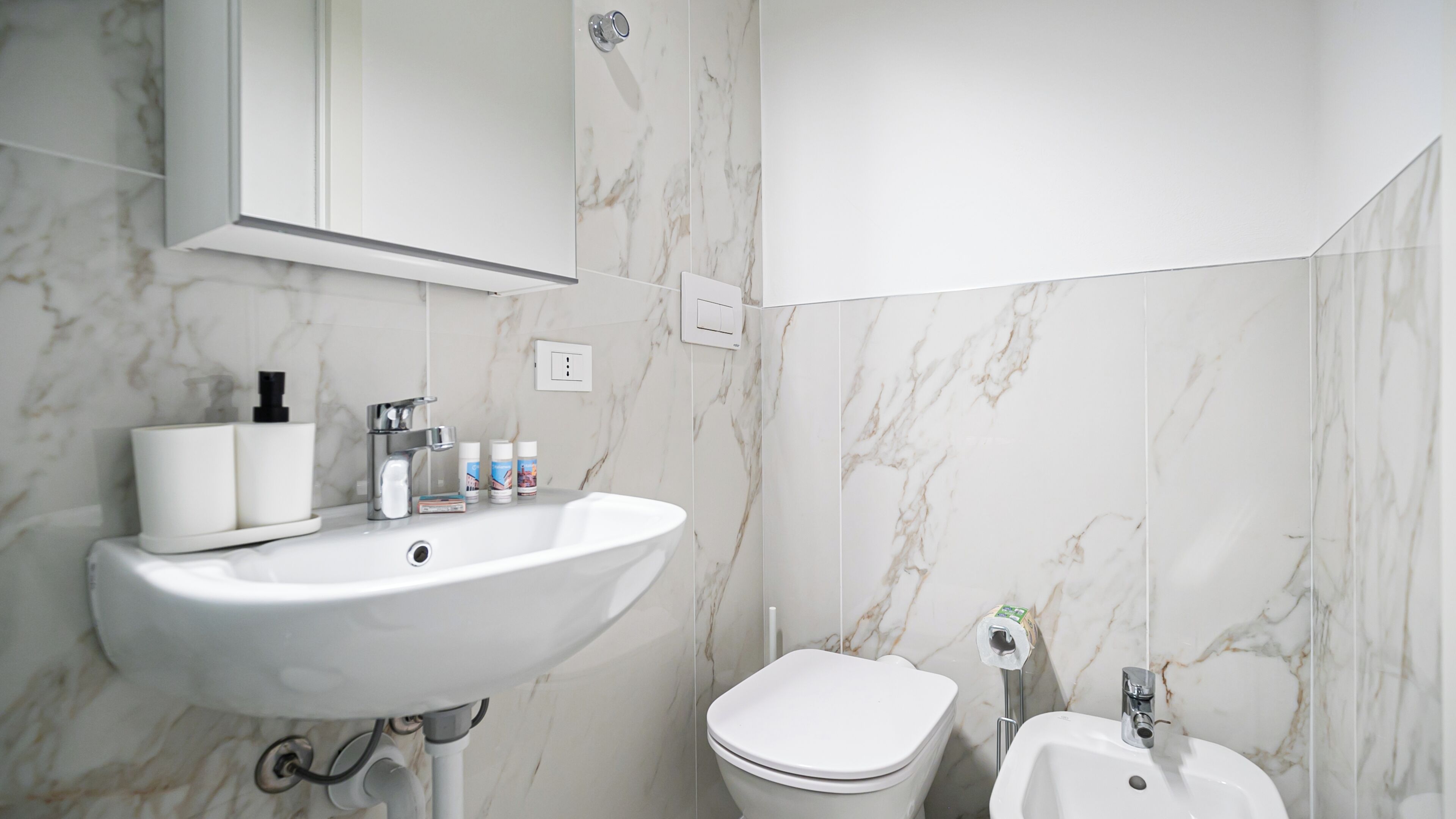 A bathroom with a white sink, toilet, and bidet, marble walls, and a mirror above the sink.