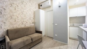 A living room with a sofa, a kitchen with white cabinets, and a grey wall. - Italianway - The B Village Standard - Apt T2 (Milan)