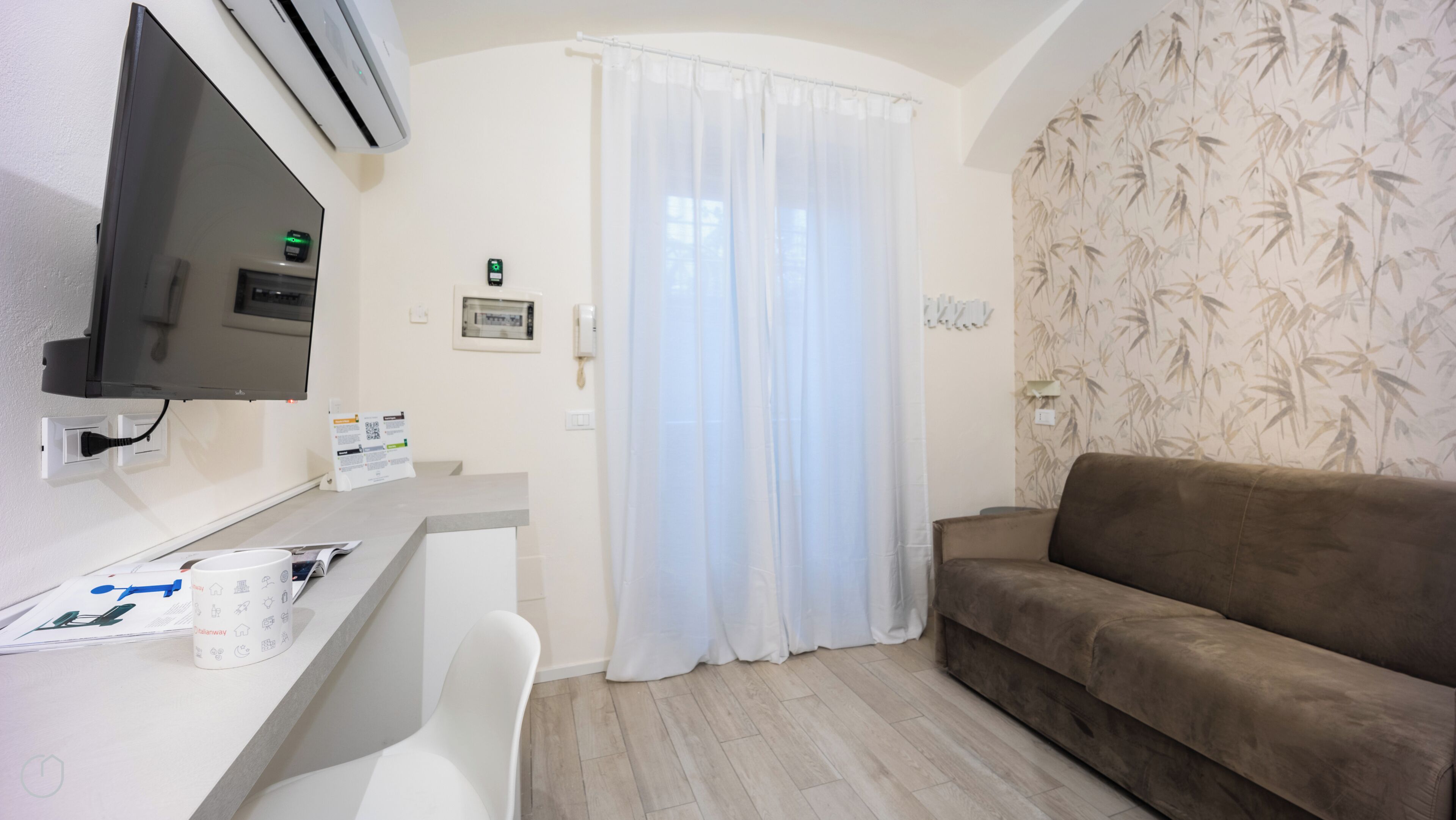 A room with a flat-screen TV, air conditioning unit, a white desk with a chair, a brown sofa, and a window with sheer curtains.