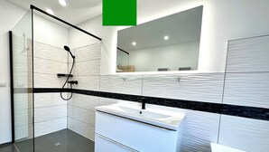 Un baño moderno con cabina de ducha de vidrio, tocador blanco con encimera negra y un espejo grande sobre el lavabo.