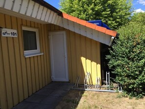 Ein gelbes Gebäude mit einer weißen Tür und einem Fenster, ein Schild mit der Aufschrift '101' und einer Metallkonstruktion zur rechten Seite.