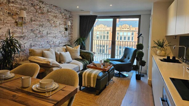 A modern living room with a brick wall, a sofa, a dining table set, and a view of buildings through large windows. - Modern Studio Greater London (New Malden)
