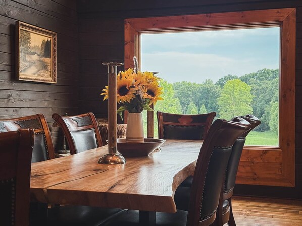 A wooden dining table with chairs, a vase with sunflowers, and a window with a view of trees.