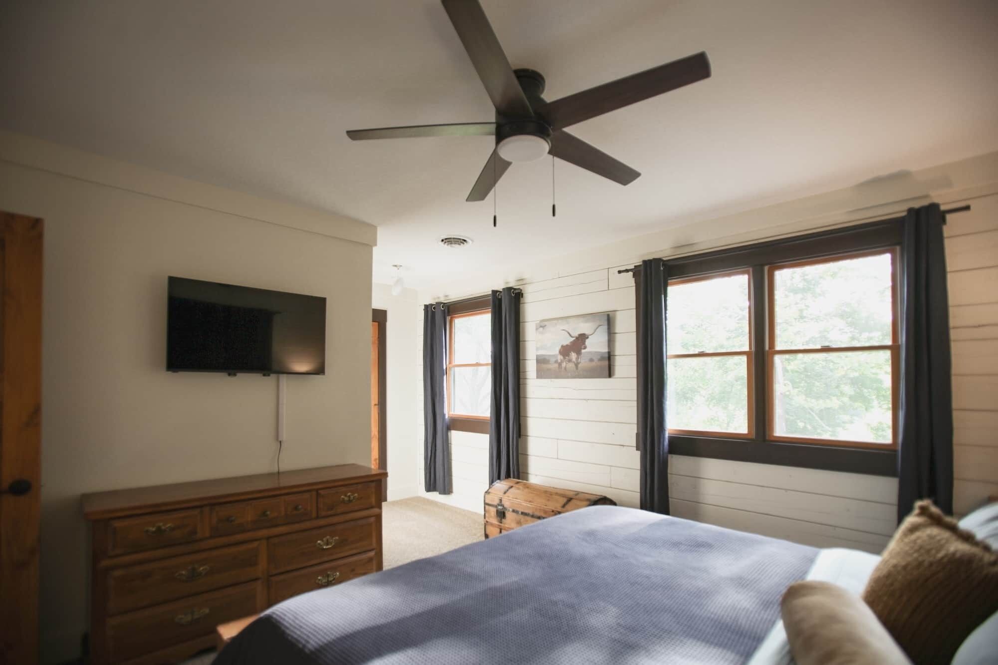 A bedroom with a bed, a dresser, a ceiling fan, a TV, and windows.