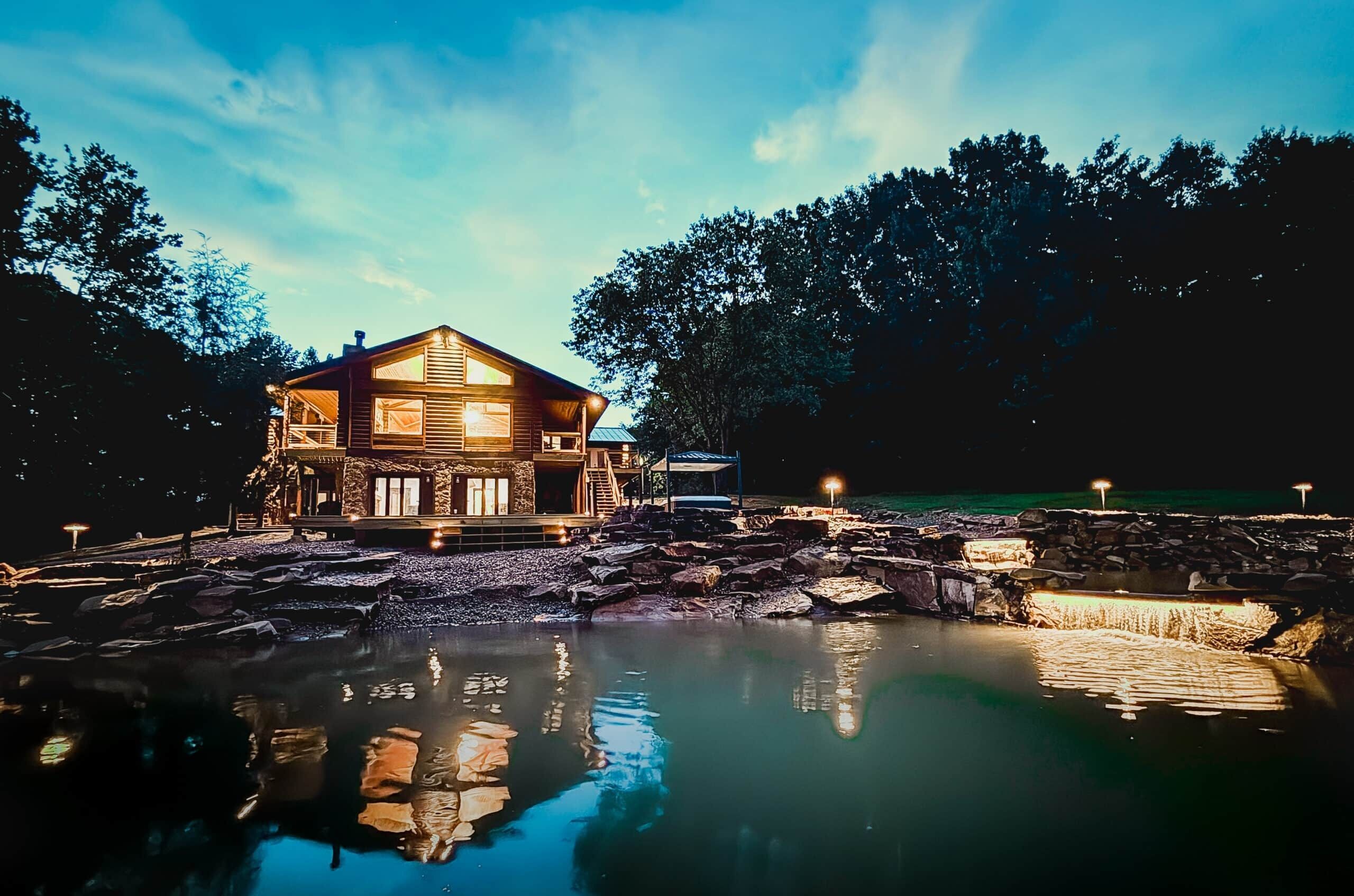 A wooden cabin with a balcony, situated by a reflective water body, surrounded by trees.