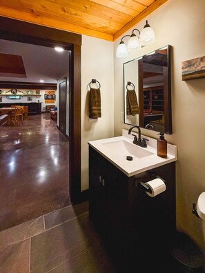 Un baño con lavamanos blanco, espejo, toallero y dispensador de papel higiénico.