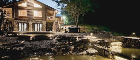 A large log cabin with a stone foundation, a wooden deck, and a pond with a stone bridge.